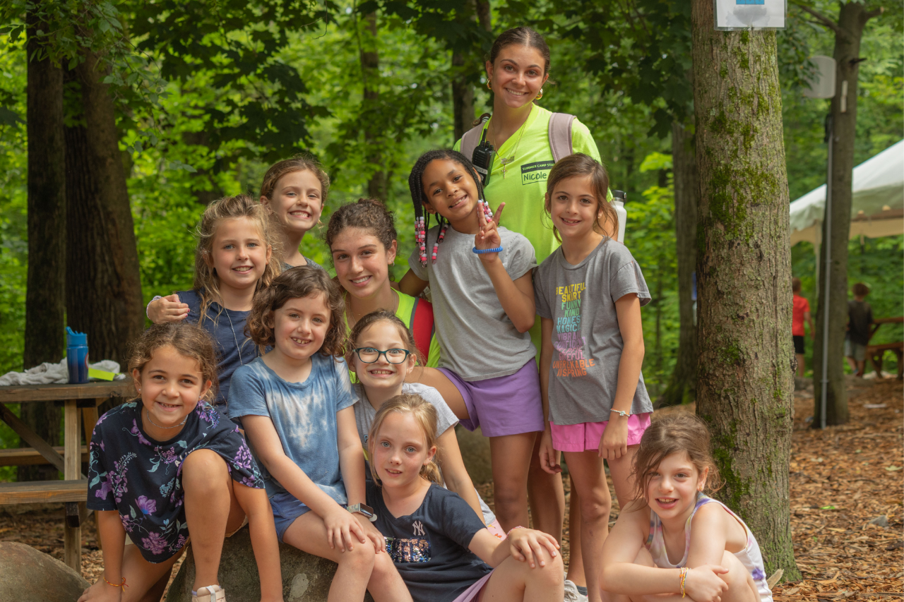 Untitled design (52) Group of children and a camp counselor posing together outdoors at a summer camp,